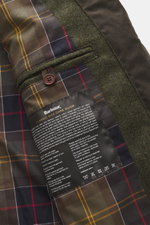 Close-up view of the interior tartan lining and care label of the Barbour Wax Jacket 'Beacon' Dark Brown, highlighting patch pockets and button strap. British style materialises in the 'Beacon' model from BARBOUR: With a slim cut and leather trim on the c