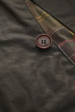 Nahaufnahme aus schräger Perspektive der dunkelbraunen Barbour Wachsjacke 'Beacon' mit gewachster Baumwolle, Tartan-Futter und Barbour-Knopf.