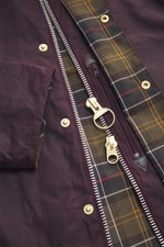 Close-up front view of the Barbour Wax jacket 'Beaufort Icons' purple showing waxed cotton fabric, silver two-way zipper, gold snap buttons, and tartan lining.