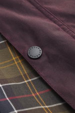 Close-up front view of the Barbour Wax jacket 'Beaufort Icons' purple showing durable waxed cotton fabric, black snap button, and characteristic tartan lining fabric.