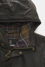 Close-up front view of Barbour Wax parka 'Bedale' dark olive showing waxed cotton hood, tartan lining, zipper, and snap buttons.

Description: An icon among wax jackets, the BARBOUR 'Bedale' returns this season as a parka, with extra length and even more 