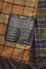 Nahaufnahme des charakteristischen Tartan-Innenfutters der Barbour Wachsjacke 'Gamefair' dunkelgrün mit Pflegehinweisen auf gewachster Baumwolle.