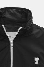 Ami Paris Blouson 'Windbreaker' schwarz, Nahaufnahme von oben, hochwertige Microfaser, glatter Griff, Regular Fit, robuster weißer Reißverschluss, im Kragen versteckte Kapuze, paspelierte Fronttaschen mit Knopf, Logo-Detail auf der Brust, elastische Saum-
