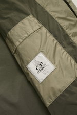 Close-up front view of C.P. Company Down jacket olive showing ripstop microfiber, textured feel, warm down filling, slim fit, garment dyed fabric, durable two-way zipper, hood with drawstring, zippered front pockets, patch arm pocket with lens detail, ela