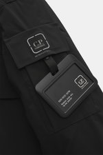 Close-up of the black C.P. Company Parka 'Long Jacket' showing the sleeve pocket with logo label and attached information tag, shot from a top-down angle.