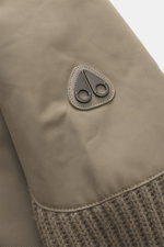Close-up detail of the typical metal logo on the upper arm of the Moose Knuckles Down blouson 'Magnus Knit Hybrid' taupe, photographed from a top side angle, featuring high-quality microfiber, knitted wool sleeves, soft touch, slim fit, durable two-way zi