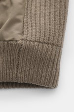 Close-up image from a side angle of the Moose Knuckles Down blouson 'Magnus Knit Hybrid' taupe, showing high-quality microfiber, knitted wool sleeves, soft touch, slim fit, durable two-way zipper, high closing ribbed collar, typical metal logo on upper ar
