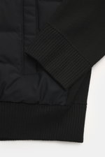 Close-up view of the Moose Knuckles Down Blouson "Kamet Hybrid" black, showing knitted back and sleeves, ribbed hem, and soft-touch viscose and microfiber fabric with light down filling. Features a robust two-way zipper, high closing ribbed collar, typica