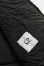 Close-up detail of the C.P. Company Daunenweste 'D.D. Shell' dark navy, showing the textured ripstop microfiber fabric and stitched brand patch, photographed from a top-down angle. Die Konstruktion der Daunenweste 'D.D. Shell' von C.P. COMPANY zeichnet si
