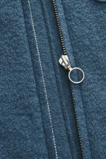 Nahaufnahme der metallischen Reißverschluss-Ziehgriffes an der graublauen 04651/ A trip in a bag Woll-Weste 'Boiled' graublau aus Bouclé-Strick, weichem Griff, Slim Fit, Stehkragen, Raw-Edge-Finish, gewalkter Schurwolle, aufgesetzter Brusttasche, kleinem 
