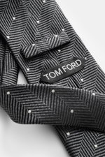 Close-up angled view of Tom Ford Silk tie dark grey/black/cream patterned, showing herringbone with polka dots, logo stitching, and silk texture.  

Description:  
<p>Confident elegance down to the finest detail: This fine silk tie by TOM FORD is a stylis