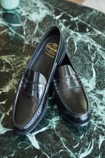 Church's Penny Loafer 'Rowley' schwarz aus poliertem Glattleder mit Ziernähten, Penny-Strap und Blick von schräg oben auf grüner Marmorfläche.