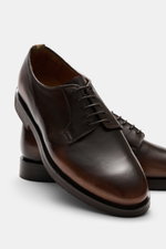 Dark taupe Officine Creative Sneaker 'Sequel' photographed from a high front angle, showcasing fine velours leather, round toe, side perforations, seven-eyelet lace-up, padded collar, logo embossing on tongue, grosgrain pull tab, leather lining, and flexi