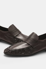 The Row Loafer dark brown shown from a slightly elevated front-side view, handcrafted in Italy from soft eel leather with a typical strap, grained look, narrowing silhouette, decorative seam on the front cap, leather lining, leather sole, and rubber inser