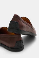 Nahaufnahme von Tod's Penny Loafer braun aus geschmeidigem Leder in schmal zulaufender Silhouette, mit Penny Strap, Steppnaht und Gummisohle mit Noppen, seitlich hinten fotografiert.