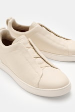 Close-up front-side view of Zegna Slip-on Sneaker 'Triple Stitch' sand in grained leather with crossed elastic bands, round toe, padded collar, pull tab, embossed tongue, leather lining, and flexible rubber sole.