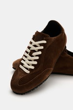 Dark brown Axel Arigato Sneaker 'Daze Runner' photographed from a close-up front angle, showing supple suede leather, seven-eyelet lacing, rounded toe cap, tonal overlays, padded collar, terry lining, and flexible rubber sole. Minimalism in motion: The 'D