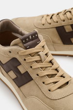 Close-up view of the Hogan Sneaker 'Allacciato' taupe/dark brown showing suede leather, seven-eyelet lacing, logo patch on tongue, high padded collar, H-shaped brand initial, leather lining, memory foam footbed, and flexible rubber sole, photographed from