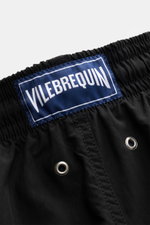 Nahaufnahme der schwarzen Vilebrequin Badeshorts 'Moorea' von hinten, zeigt elastischen Bund, Tunnelzug, Entwässerungsösen und das blaue VILEBREQUIN-Weblabel, gefertigt aus hochwertiger Microfaser mit leichtem Griff und Mesh-Innenfutter, ideal für Sommer,