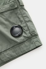 Nahaufnahme der graugrünen C.P. Company Cargo-Bermudas, schräg von oben fotografiert, mit Label-Linsendetail und Cargo-Tasche.