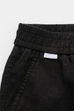 Nahaufnahme von 04651/ A trip in a bag Leinenshorts schwarz mit elastischem Bund, französischer Tasche und kleinem Weblabel, aus luftigem Leinen, weicher Griff, garment dyed, Regular Fit, aufgesetzte Gesäßtasche, frontal fotografiert.