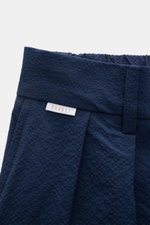 Nahaufnahme der Taille der 04651/ A trip in a bag Seersucker-Shorts navy aus strukturtypischem Microfaser-Seersucker mit teilelastischem Bund, doppelter Bundfalte und kleinem Weblabel, fotografiert von schräg vorne.
