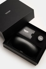 Das Bild zeigt aus schräger Draufsicht eine geöffnete schwarze Geschenkbox mit Leder-Hartschalenetui, Horn-Politurset und Putztuch der Tom Ford Horn Brille 'Private Collection' schwarz/graubraun/weiß gemustert.