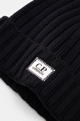 Close-up image taken from an angled top-down perspective showing the cuff with logo patch of the C.P. Company Merino beanie dark navy featuring pure merino wool, soft feel, classic ribbed knit pattern, and medium coarse knit texture.