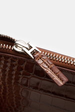 Nahaufnahme einer braunen Tom Ford Tasche mit Reptilien-Leder-Optik, silberfarbenem Messing-Reißverschluss und Logo-Prägung.