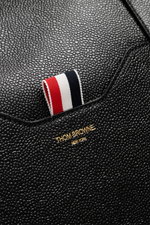 Close-up of the Thom Browne Weekender schwarz showing finely grained black leather, tricolor fabric tab, and gold logo from a top-front angle, highlighting luxurious texture and craftsmanship. <p>Bereit für den nächsten Kurztrip: Der geräumige Weekender v