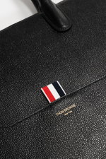 Close-up top view of the Thom Browne Shopper schwarz showing finely grained black leather, sturdy handle, signature tricolor ribbon tab, and gold logo. Souveräner Stil für jeden Tag: Der geräumige Shopper von THOM BROWNE aus fein genarbtem Leder vereint e