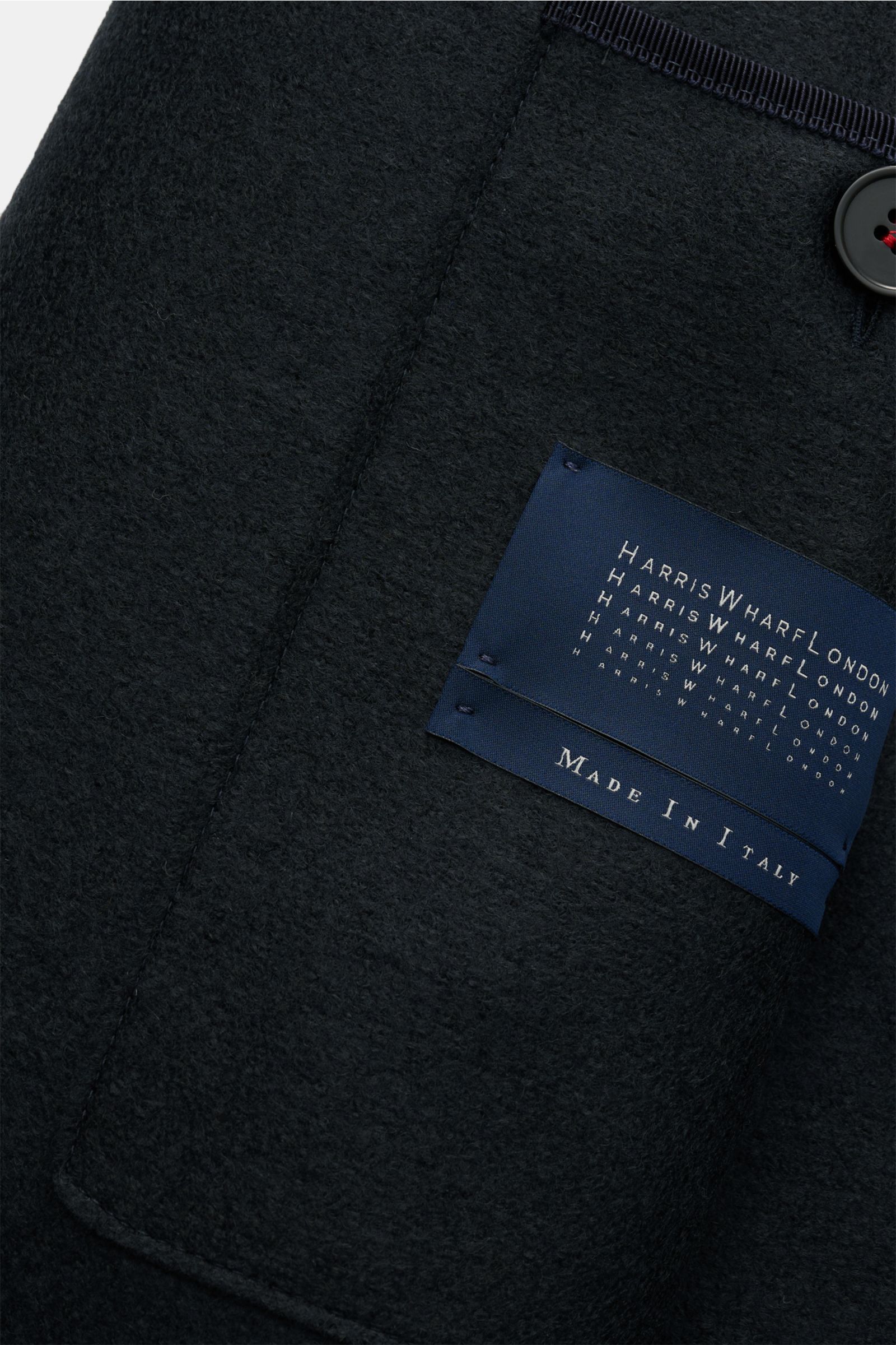 Close-up angled view of the Harris Wharf London Wollmantel navy label on heavy, felted virgin wool fabric with a button and stitched edges, showing texture and craftsmanship. Zeitlos, pur und durchdacht: Dieser ungefütterte Mantel mit gerader Silhouett