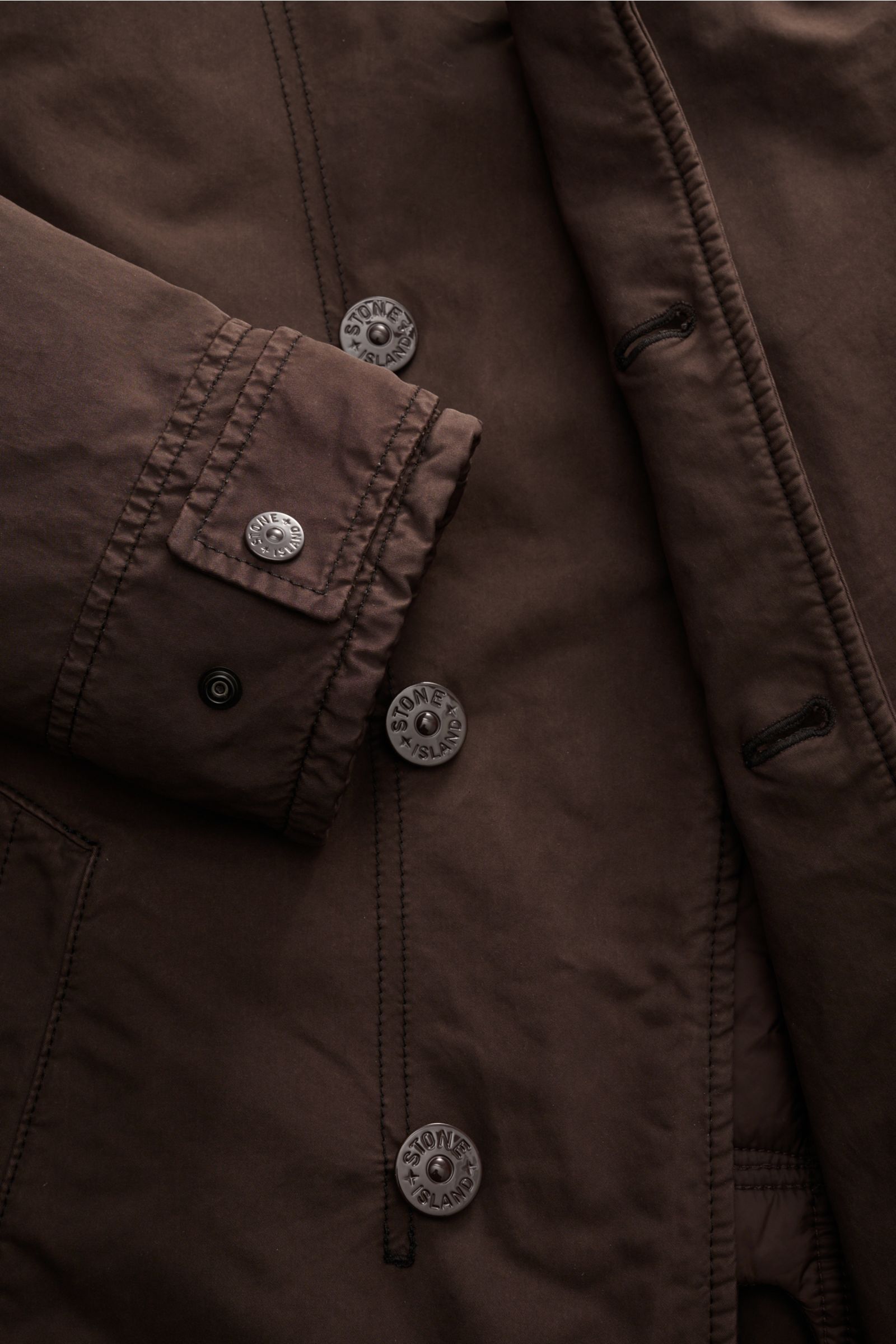 Nahaufnahme der Stone Island Jacke 'David-TC Ghost Piece' dunkelbraun, Front mit Knöpfen, Manschette und tonalem Badge sichtbar.