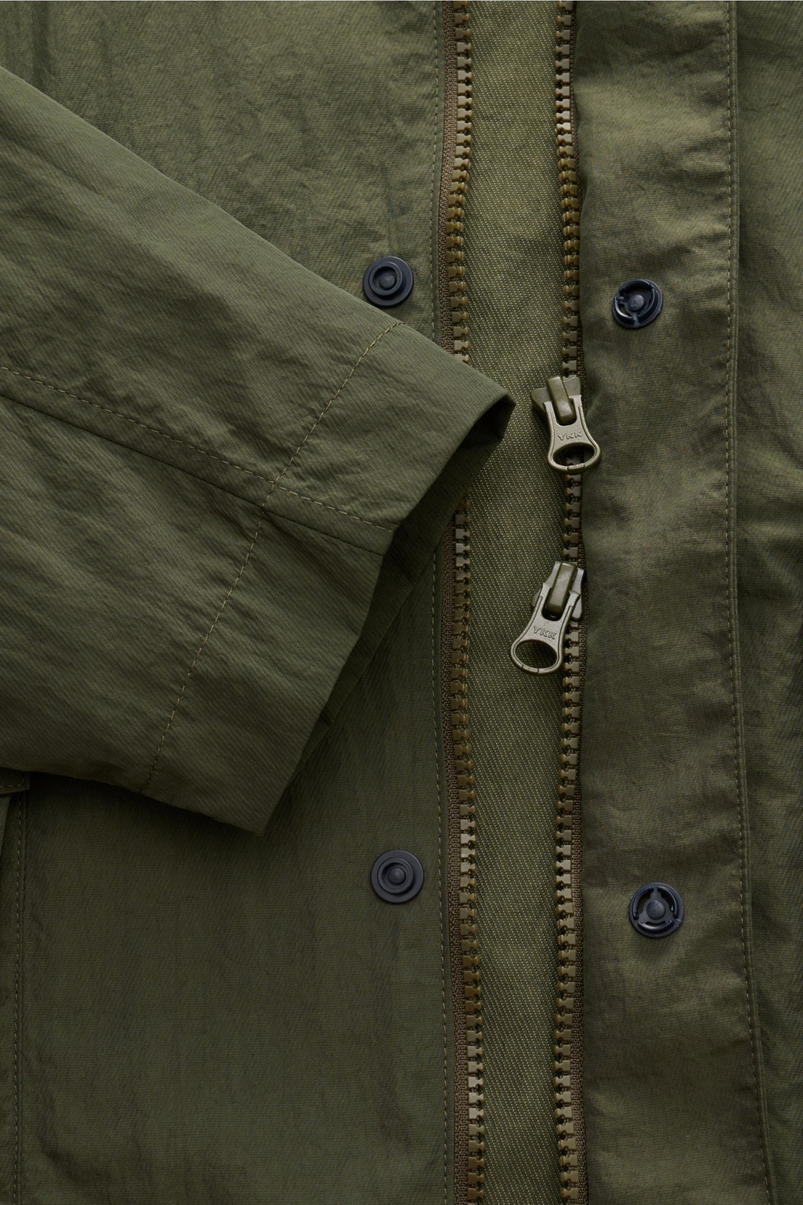 Alt-Tag (124 Zeichen): Detailaufnahme des olivfarbenen 04651/ A trip in a bag Mantel 'Travel Coat' mit Reißverschluss, Druckknöpfen und Microfaser.