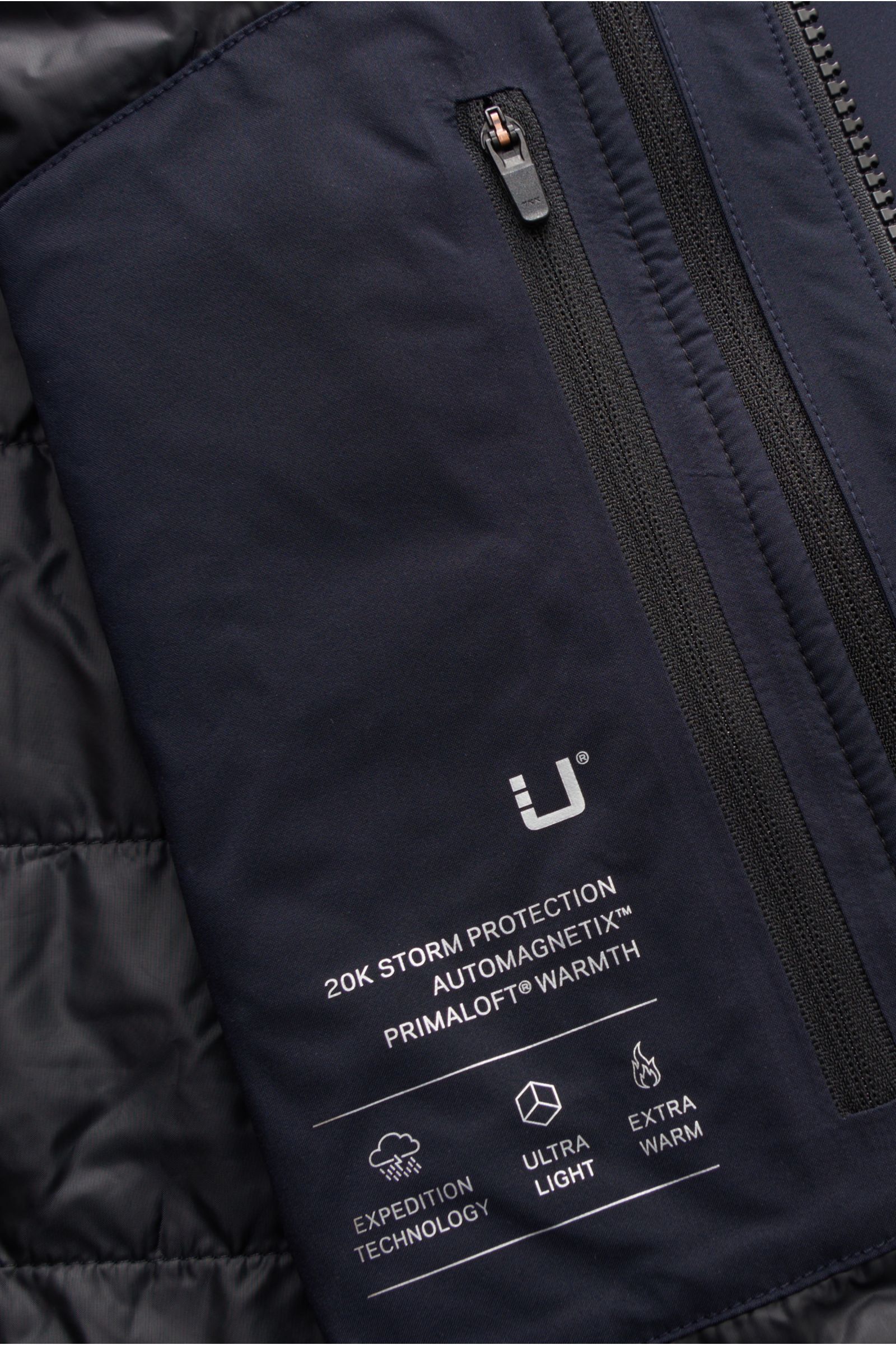 Close-up front interior view of the UBR Mantel 'Black Storm' navy showing technical microfiber fabric with printed logos and a zippered pocket. Dank High-Tech trocken durch Regen und Sturm: Der cleane 'Black Storm' Mantel von UBR aus technischer Microfase