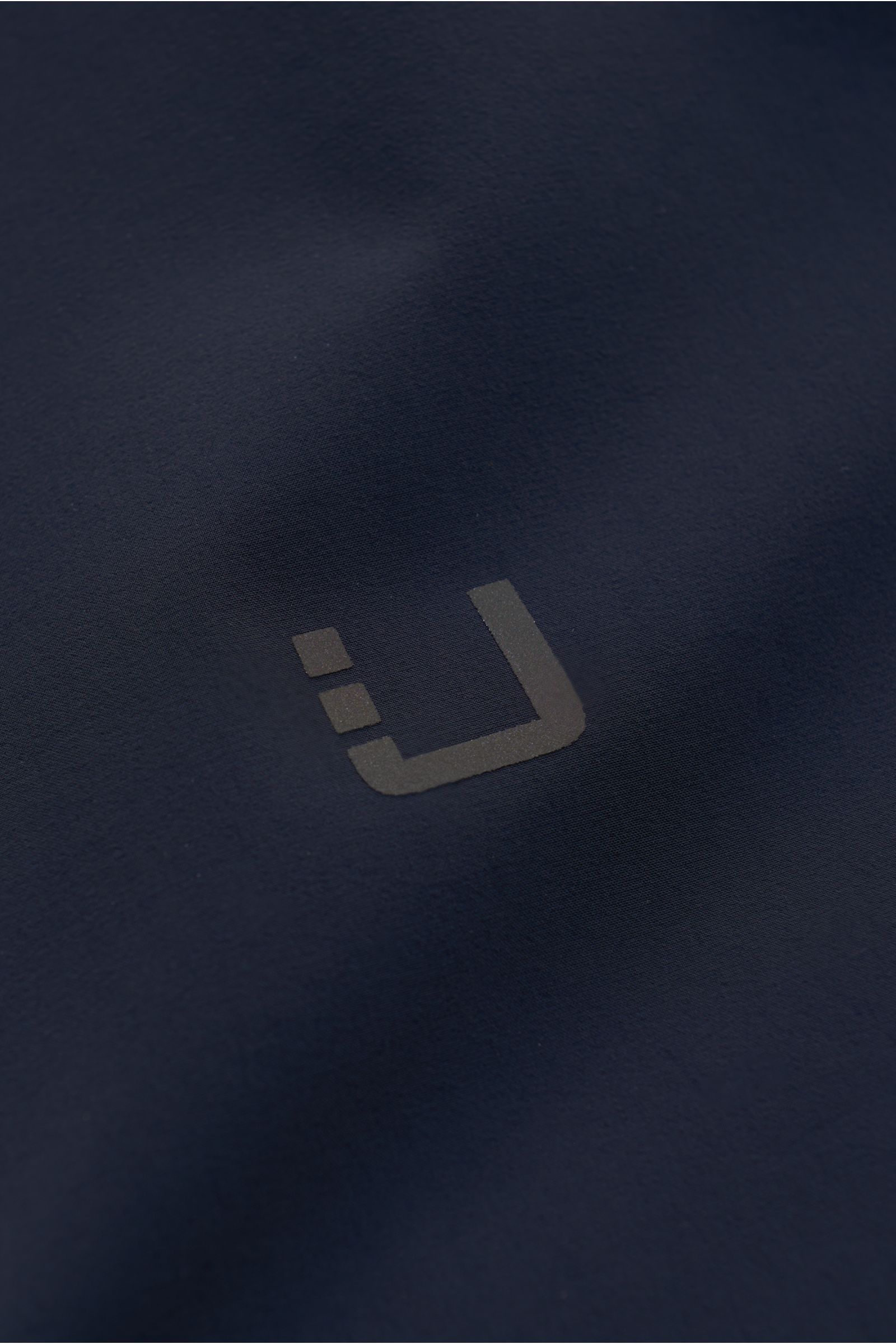 Close-up front view of the reflective logo on the navy fabric of the UBR Kurzmantel 'Regulator Coat', showcasing its smooth, high-quality microfiber material with bi-elastic and water-resistant properties.