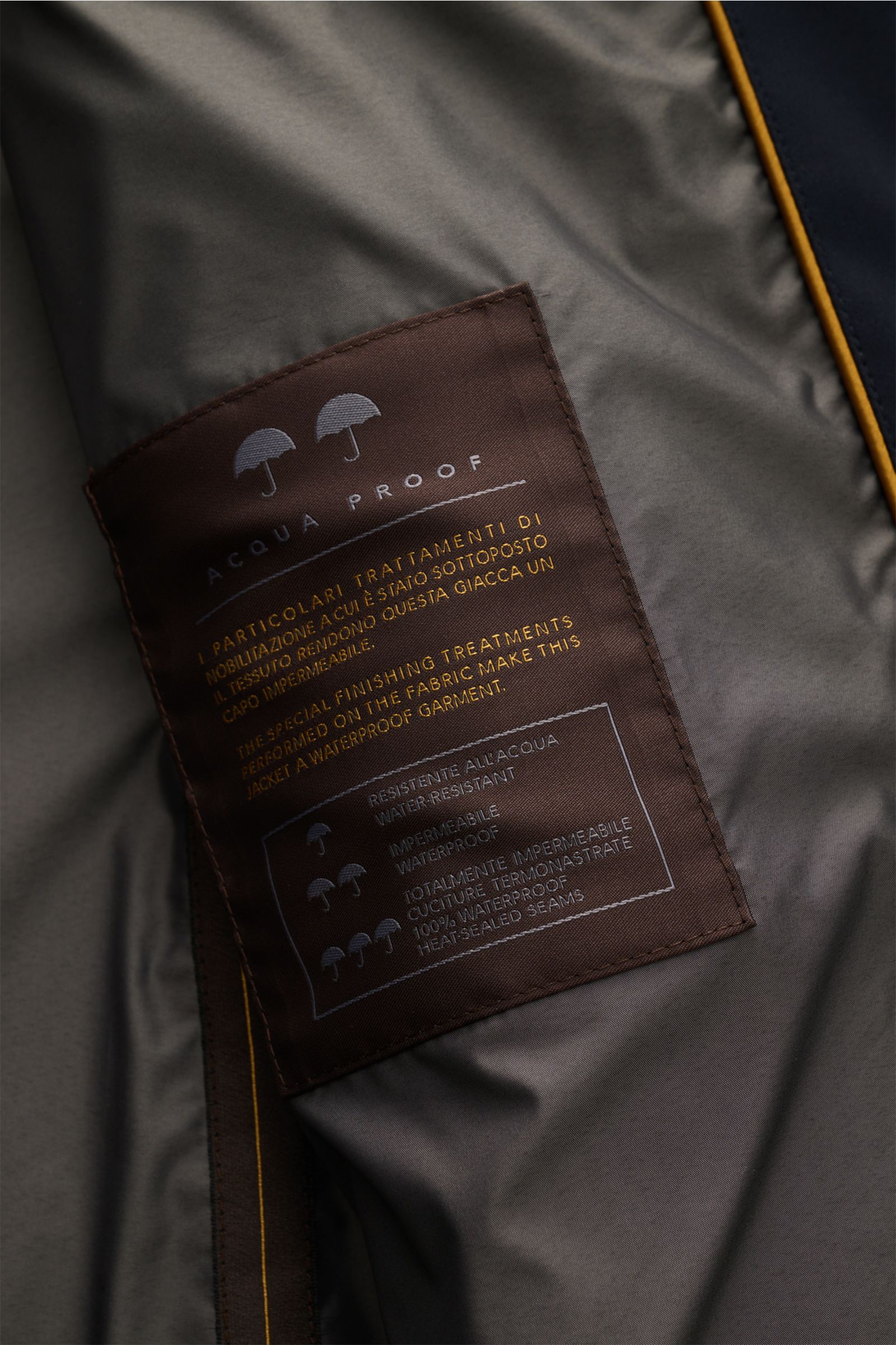 Close-up inside view of the Moorer Kurzmantel 'Borromini' navy showing the brown waterproof fabric label with care instructions and waterproofing details, photographed from above. Eleganter Begleiter für wechselhaftes Wetter: Der Kurzmantel 'Borromini' vo