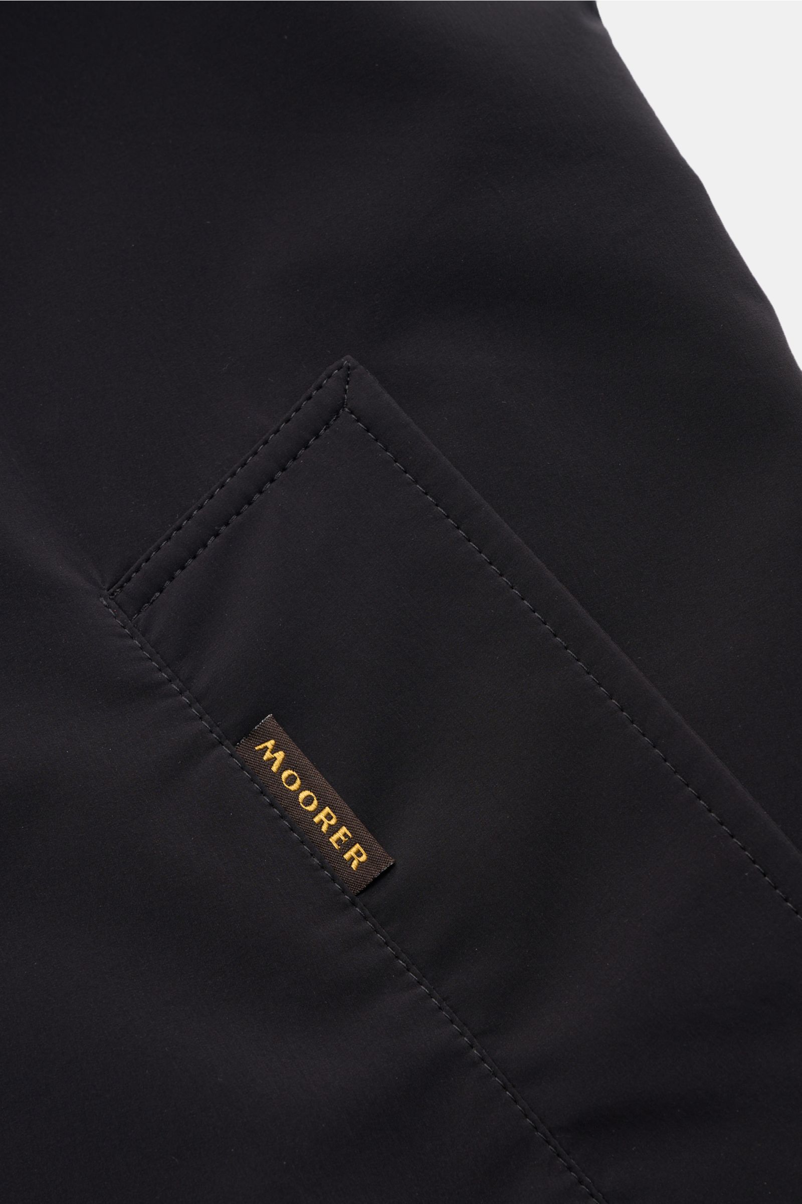 Close-up angled view of the Moorer Kurzmantel 'Vittor' navy pocket with Moorer label, water-repellent microfiber, slim fit, four-button front, turn-down collar, piped front pockets, buttoned cuffs, back vent, smooth lining, and inner pocket.