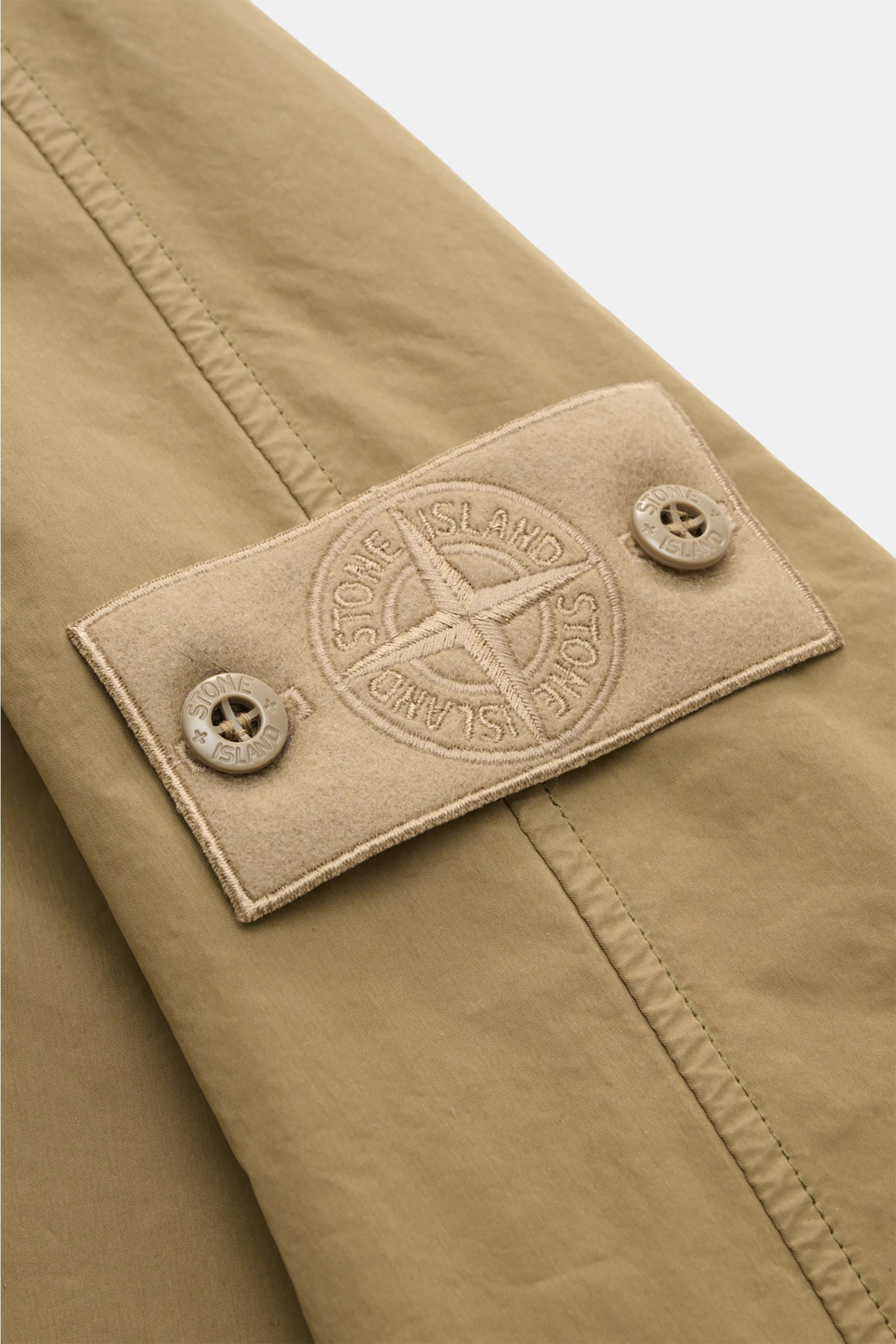 Close-up side view of the tonal Stone Island Coat 'Ghost Piece' khaki sleeve featuring the embroidered compass badge on a lightweight, water-repellent fabric.