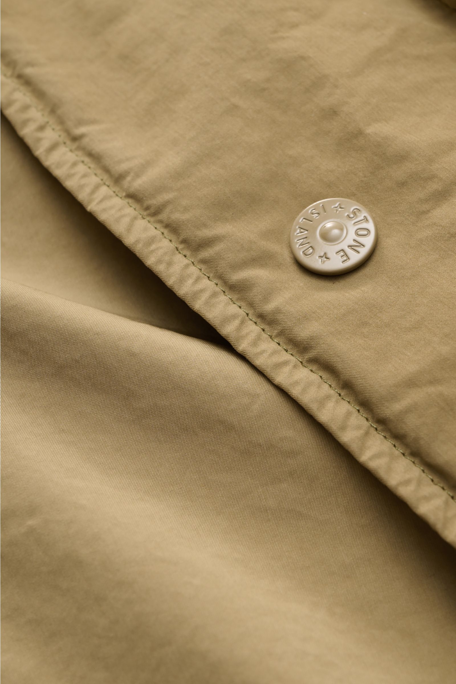 Close-up front view of the Stone Island Coat 'Ghost Piece' khaki showing its high-quality microfibre fabric, light velvety feel, and branded snap button. In the tonal design of the 'Ghost' collection, this lightweight urban coat by STONE ISLAND in 'David 