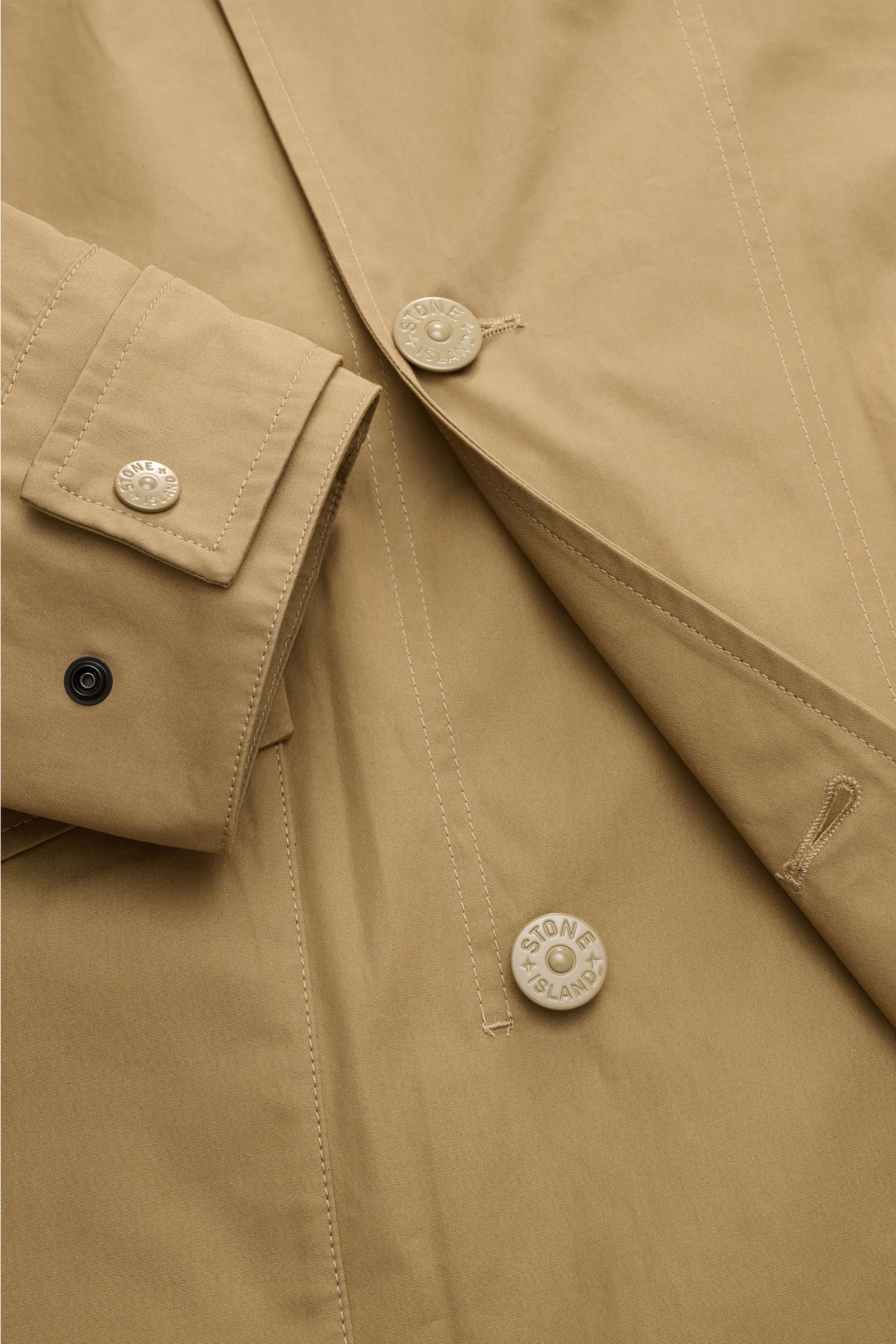 Close-up top-down view of Stone Island Kurzmantel 'Ghost Piece' khaki showing water-repellent popeline, two-row three-button placket, and adjustable cuff with tonal buttons.

Eleganz trifft Funktion: Der Kurzmantel von STONE ISLAND aus der 'Ghost-Colle