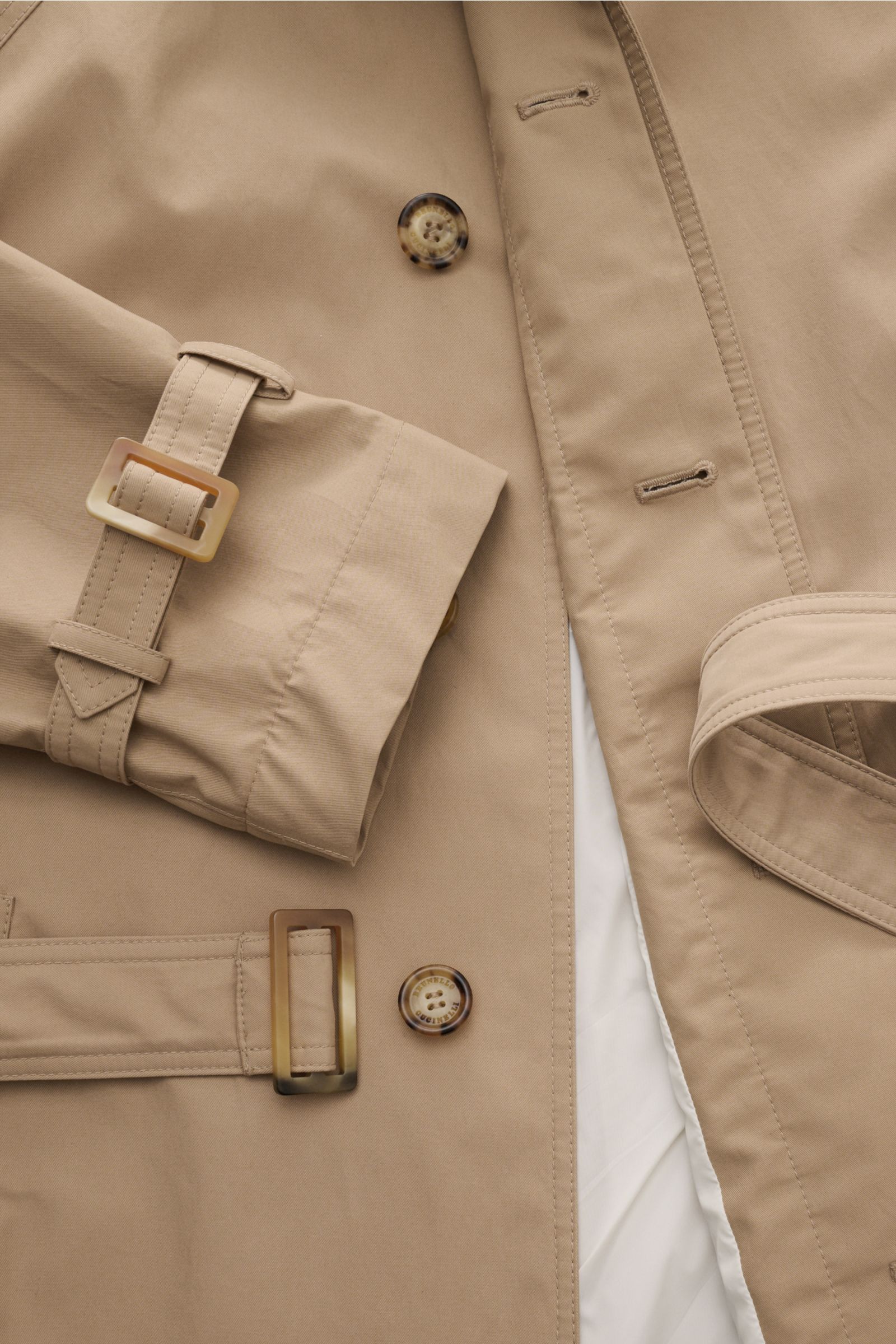 Close-up front view of beige Brunello Cucinelli Trenchcoat showing button details, belt with horn buckle, cuff strap, and smooth fabric texture. Wasserabweisend und atmungsaktiv: Ein innovativer Mix aus Baumwolle und Microfaser ist die Grundlage für diese