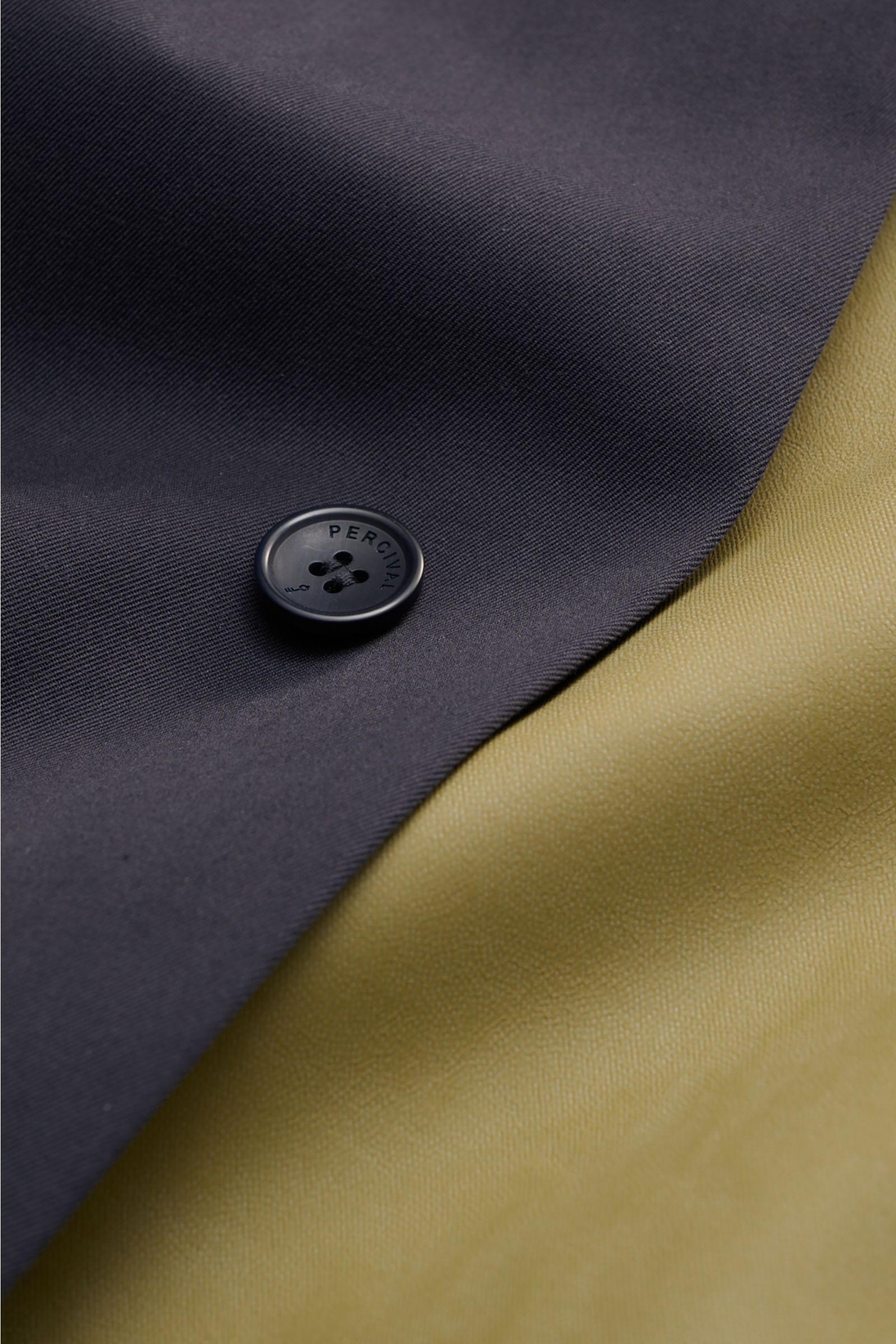 Close-up top view of the Percival Kurzmantel 'Sherlock' dark navy showing durable twill fabric with a branded button, next to light olive lining; the coat features a mix of cotton and microfiber, waterproof with a 10k/10k rating, sealed seams, C0-DWR eco-