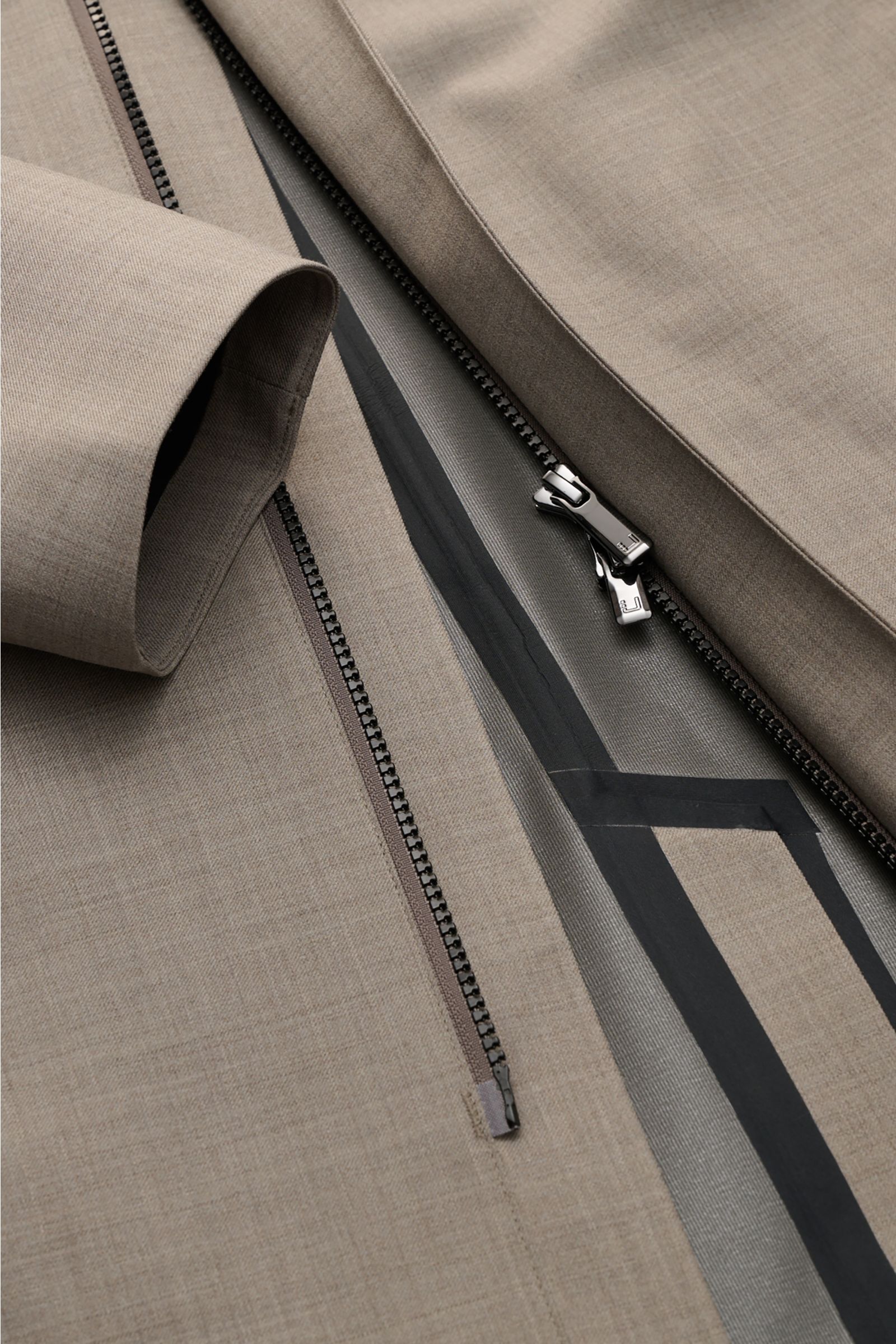 Close-up, top-down view of the zipper area of the UBR Rain coat 'Sky Fall Savile' taupe mottled, showing taupe mottled fabric with black waterproof zippers and taped seams. 

The incredibly lightweight rain coat 'Sky Fall Savile' by UBR is made of smar