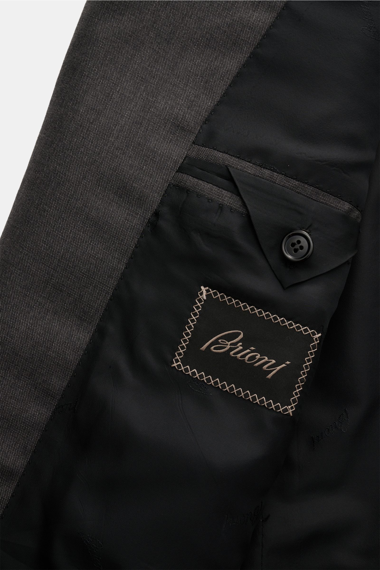 Close-up image of the inside lining and fabric texture of the Brioni Anzug 'Brunico' dunkelgrau meliert, showing the tailored black satin interior with branded stitching and buttoned pocket, photographed from an angled top-down perspective. Elegance from 