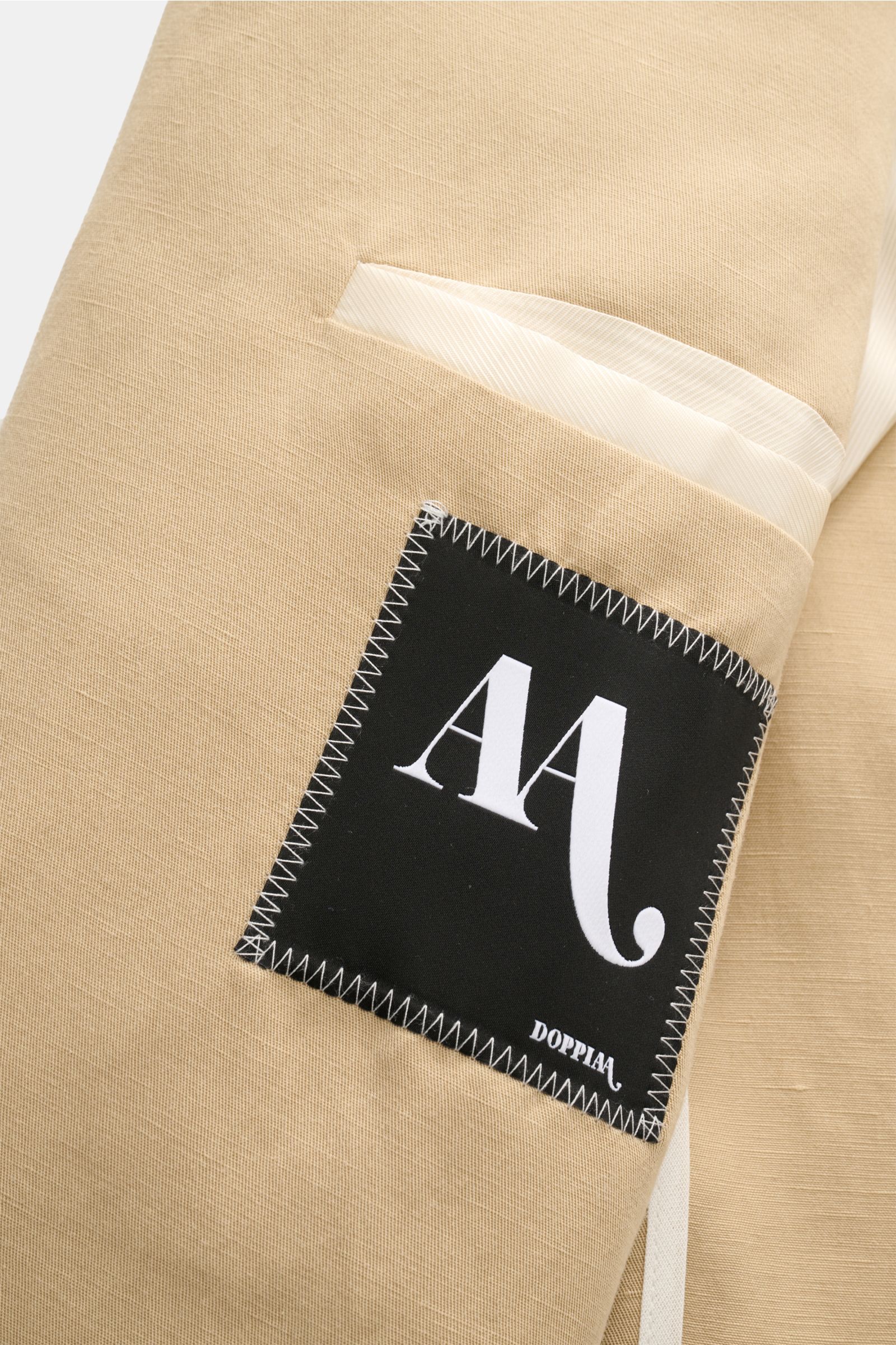 Close-up of the beige Doppiaa Anzug 'Aanetoant' showing the fabric texture and stitched black label, photographed from an angled top view.