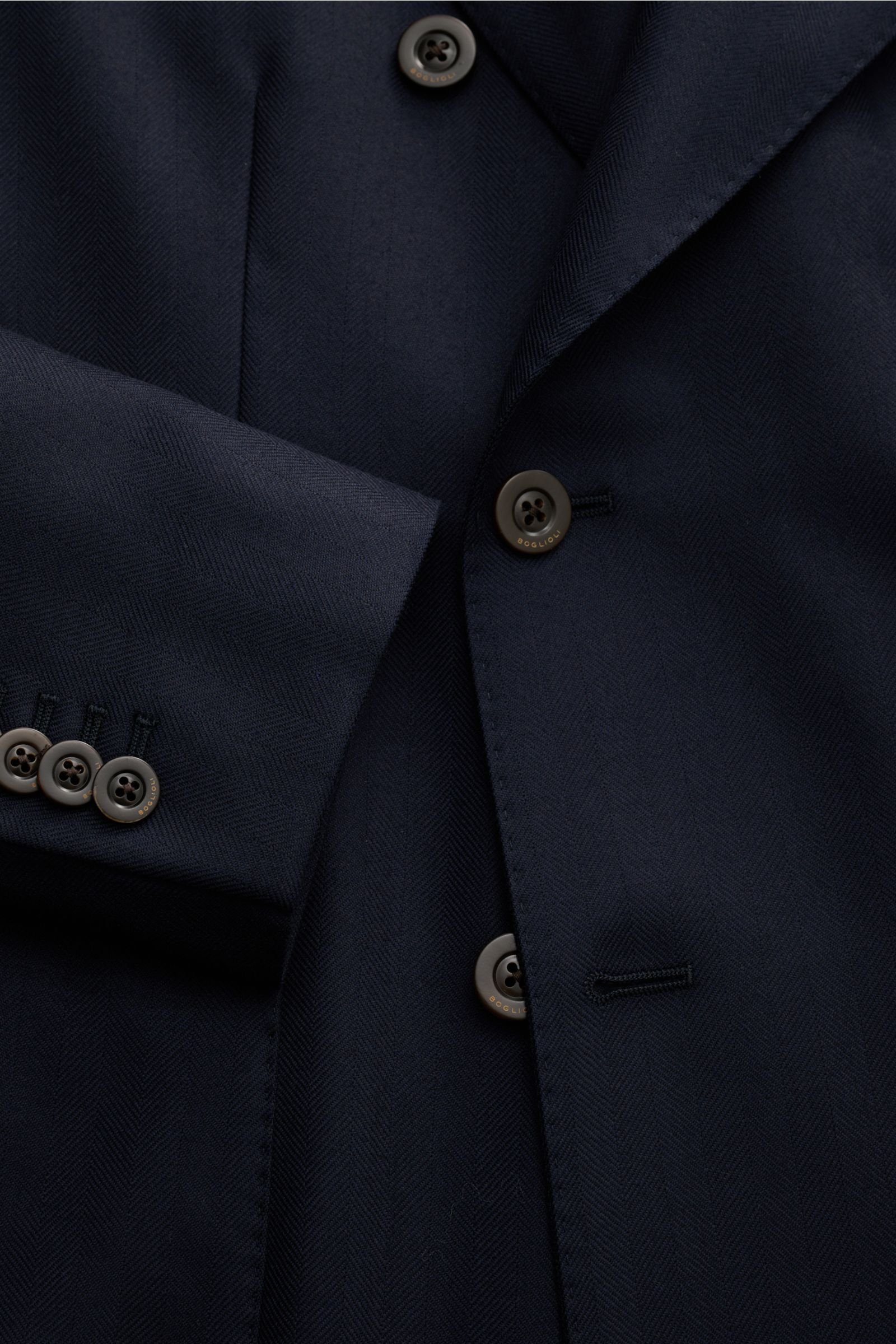 Close-up view of the Boglioli Sakko 'K. Jacket' navy showing tonal herringbone pure virgin wool fabric, slim fit, two-row three-button style, peaked lapel, classic breast pocket, patch hip pockets, kissing buttons, rear side vents, unconstructed design, a