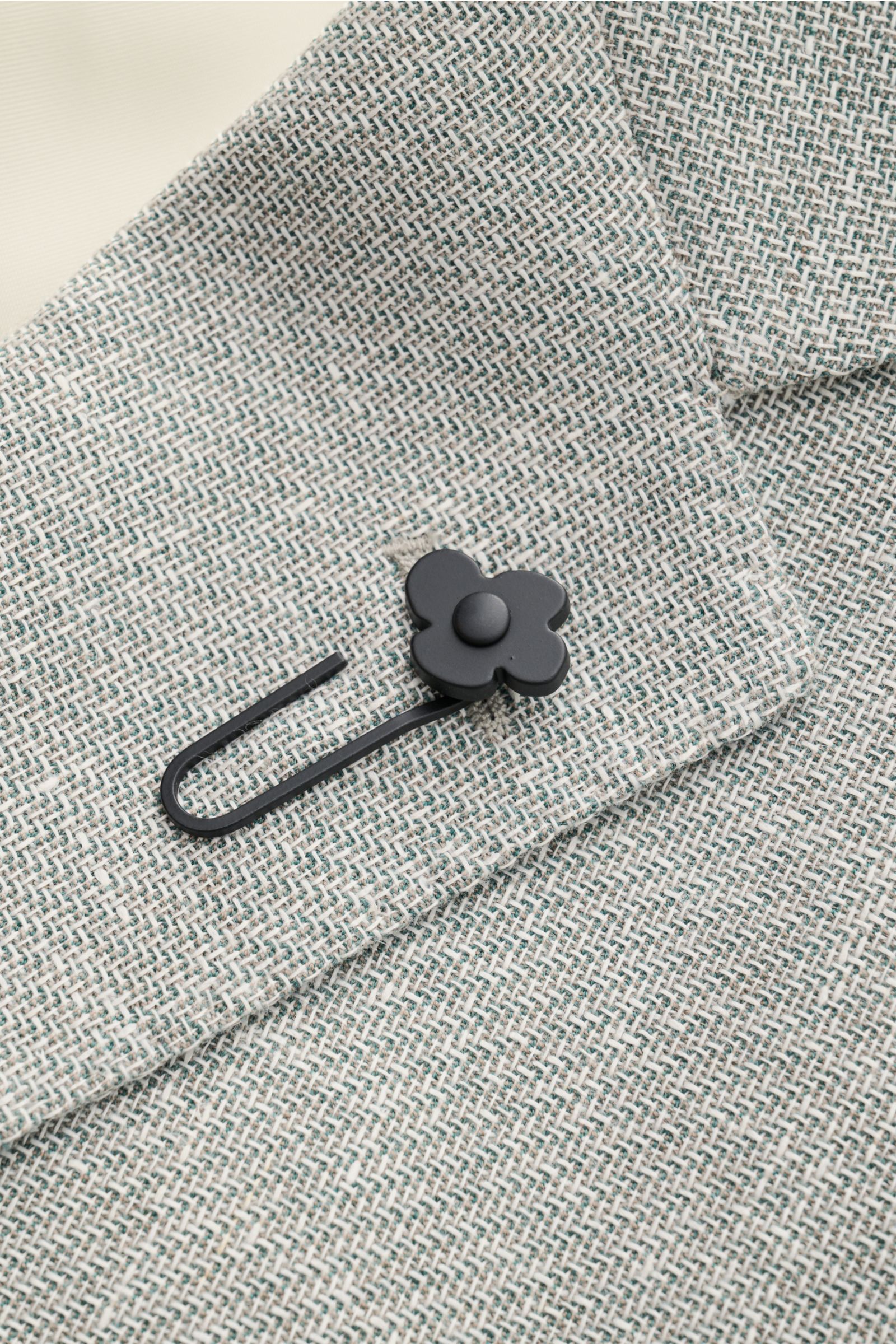 Nahaufnahme aus schräger Draufsicht des Lardini Sakko salbei/offwhite meliert mit strukturierter Schurwolle-Leinen-Webstruktur und schwarzem Blumen-Anstecker.