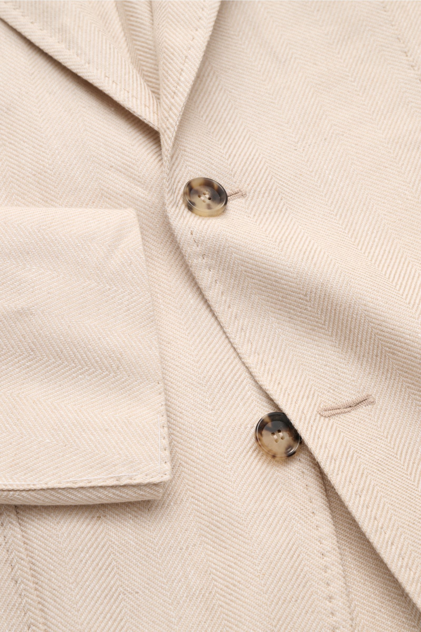 Close-up front detail of the Sipario Smart-casual jacket 'Ginza' sand patterned, showing tonal herringbone fabric, two buttons, step lapel, and classic breast pocket from an angled top perspective.

Description: Lightweight and stylish: The smart-casua