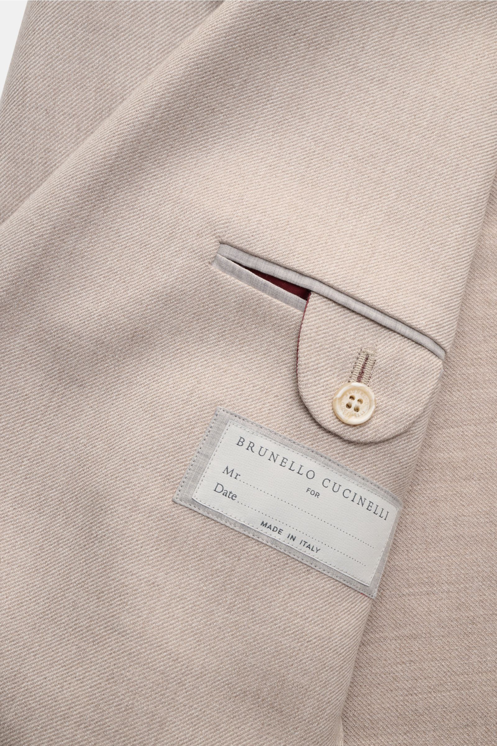 Nahaufnahme des sand melierten Brunello Cucinelli Sakkos mit markanter Webstruktur, weichem Griff und Innenetikett, seitlich fotografiert.